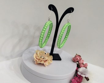 May include: A pair of green hoop earrings with a textured surface. The earrings are displayed on a black stand with a white base.