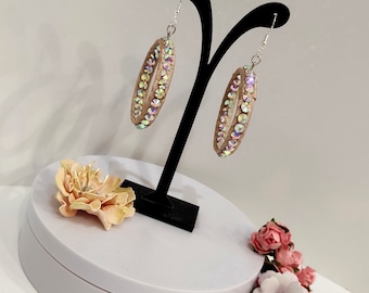 May include: A pair of hoop earrings with a beige base and iridescent rhinestones. The earrings are displayed on a black stand.