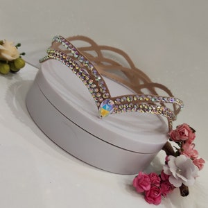 May include: A light brown headband with a decorative design of iridescent rhinestones. The headband has a V-shaped design with a large iridescent rhinestone in the center.