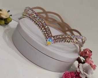 May include: A light brown headband with a decorative design of iridescent rhinestones. The headband has a V-shaped design with a large iridescent rhinestone in the center.