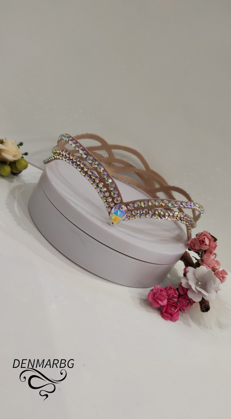 May include: A light brown headband with a decorative design of iridescent rhinestones. The headband has a V-shaped design with a large iridescent rhinestone in the center.