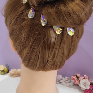 Puede incluir: Una cabeza de maniqu&iacute; con cabello casta&ntilde;o claro peinado en un mo&ntilde;o, adornada con varias horquillas de cristal iridiscente. Los alfileres tienen forma de l&aacute;grima y reflejan m&uacute;ltiples colores. El texto "DENMARBG" es visible.