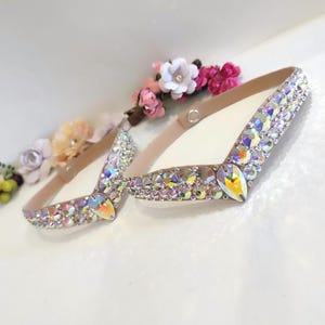 May include: Two beige elastic headbands with a V-shaped design, adorned with iridescent rhinestones and a large teardrop-shaped crystal in the center.