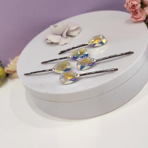 Puede incluir: Seis horquillas para el pelo con detalles de cristal iridiscente en forma de l&aacute;grima. Los cristales reflejan un arco&iacute;ris de colores. Las horquillas est&aacute;n dispuestas sobre una superficie blanca, con un fondo morado suave y detalles florales.
