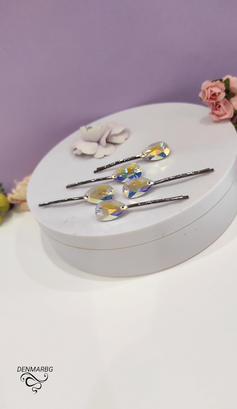 Puede incluir: Seis horquillas para el pelo con detalles de cristal iridiscente en forma de l&aacute;grima. Los cristales reflejan un arco&iacute;ris de colores. Las horquillas est&aacute;n dispuestas sobre una superficie blanca, con un fondo morado suave y detalles florales.