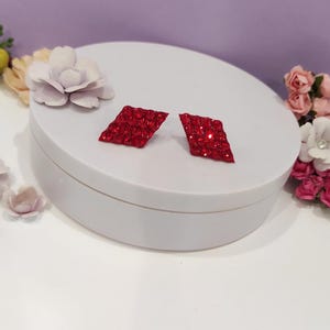 Könnte beinhalten: Zwei rote, rautenförmige Ohrringe mit glitzernder Oberfläche. Sie liegen auf einem weißen, kreisförmigen Podest, umgeben von künstlichen Blumen in Rosa, Weiß und Gelb. Der Hintergrund ist ein helles Lila. Die Ohrringe sind etwa 2 cm lang.