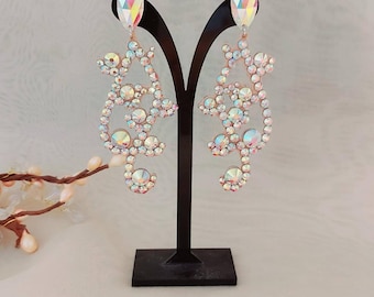 May include: A pair of iridescent crystal earrings with a floral design. The earrings are displayed on a black stand.