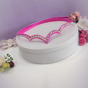 Puede incluir: Una diadema rosa con un borde festoneado y una fila de piedras de cristal iridiscentes.