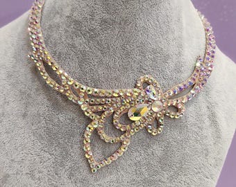 Puede incluir: Un collar iridiscente y brillante con un diseño floral, exhibido en un busto gris. El collar presenta numerosos cristales claros y coloridos, creando un efecto deslumbrante. La marca "DENMARBG" es visible.