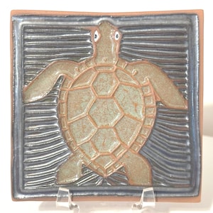 May include: Square ceramic tile featuring a sea turtle design. The turtle is in shades of green and brown, with a textured shell. The background has a blue and gray wave pattern, framed by a brown border. The tile is displayed on a clear stand.