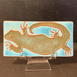May include: A rectangular ceramic tile featuring a stylized lizard design. The lizard is olive green with orange outlines, set against a light blue background. The tile is displayed on a clear acrylic stand.