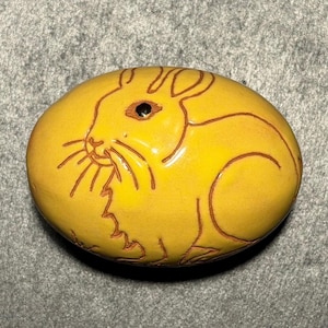 May include: A smooth, oval-shaped yellow stone featuring a dark brown etched outline of a rabbit. The rabbit is depicted in profile, showing long ears, whiskers, and a small dark eye. The surface of the stone has a glossy finish, highlighting the engraved details of the animal's form against the bright yellow background.