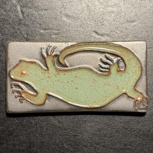 May include: A rectangular ceramic tile featuring a stylized lizard design. The lizard is light green with brown accents, set against a silver-gray background. The tile is likely handmade and suitable for home decor.
