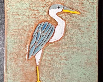 May include: A square ceramic tile featuring a raised, hand-painted relief of a heron. The bird has a grey head, a yellow beak, a white neck, and blue-toned wings. The background is a textured, light mint green with brown undertones.