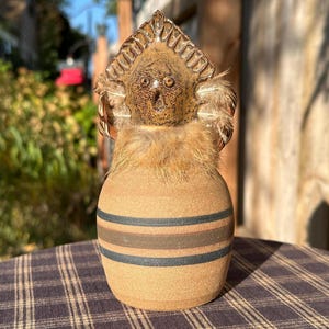 May include: A ceramic figurine with a brown and tan striped body, a brown face with large eyes, and a brown feathered headdress. The figurine is sitting on a plaid tablecloth.