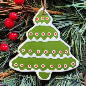 May include: A green Christmas tree ornament with white trim and red dot accents. The tree-shaped decoration is hanging from a brown string. Red berries and green pine needles are in the background.