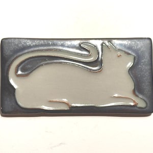 May include: A rectangular ceramic tile featuring a stylized cat design. The cat is light gray, with a curled tail and a raised head, set against a dark gray background. The tile has a glossy finish and a raised relief design.