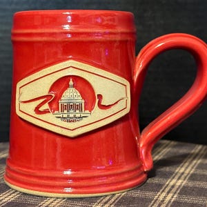 May include: A red ceramic beer stein with a beige handle and a beige emblem featuring a building with a dome and a gold ribbon.