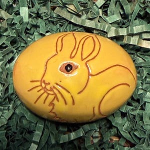May include: A hand-painted, oval-shaped stone with a yellow base and a brown outline of a rabbit. The rabbit has a brown eye and whiskers. The stone is on a bed of green shredded paper.