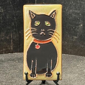 May include: A rectangular ceramic tile featuring a black cat with green eyes, a red collar, and a red pendant. The cat is set against a gold-colored background. The tile is displayed on a black metal stand.