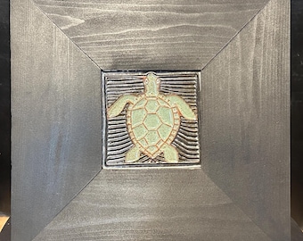 May include: Square wooden framed artwork featuring a ceramic turtle. The turtle is light green with a brown outline, set against a textured background. The frame is dark gray, and the artwork is displayed on a black stand.