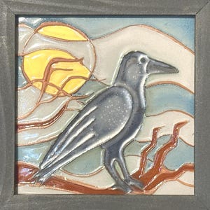 May include: Square ceramic tile with a framed design featuring a gray crow perched on a branch. The background includes a yellow sun and a blue and white sky. The tile is framed in a gray wooden frame.