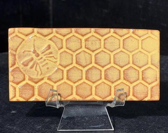 May include: A rectangular ceramic tile featuring a raised honeycomb pattern in shades of yellow and orange. A circular embossed design of a bee is positioned on the left side of the tile. The tile is displayed on a clear plastic stand.