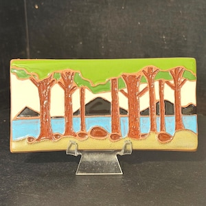 May include: A rectangular ceramic tile featuring a landscape scene with brown tree trunks, a bright green canopy, a light blue body of water, and dark brown mountains against a cream background, all resting on a tan foreground.