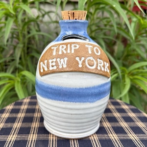 May include: White ceramic piggy bank with blue stripes and a brown label that reads "Trip to New York".