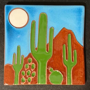 May include: A square ceramic tile featuring a desert landscape with a bright blue sky, a white sun, brown mountains, and several green saguaro and prickly pear cacti. The design is rendered in a glossy, raised relief style with distinct color sections.
