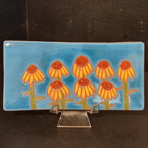 May include: A rectangular ceramic tray with a glossy blue background featuring a hand-painted design of eight yellow coneflowers with orange centers and green stems. The tray is displayed on a clear plastic stand, showcasing the vibrant floral art.
