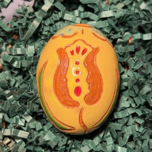 May include: A hand-painted, yellow Easter egg with orange and red floral designs. The egg is decorated with a tulip-like pattern and green accents. The egg is nestled in green paper filler.