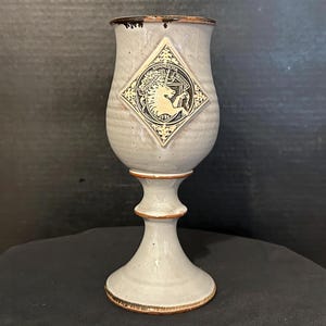 May include: A gray ceramic goblet with a pedestal base and a flared rim. A diamond-shaped emblem featuring a unicorn and the words "University of Minnesota" is centered on the goblet. The rim and base have a brown trim.