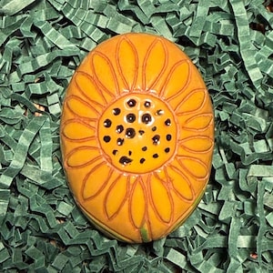May include: A hand-painted, oval-shaped ceramic sunflower with yellow petals and a dark center. The sunflower is set against a background of green shredded paper. The design features detailed lines and a glossy finish.