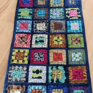 May include: A blue quilted table runner with a patchwork design featuring various colourful squares with geometric patterns.