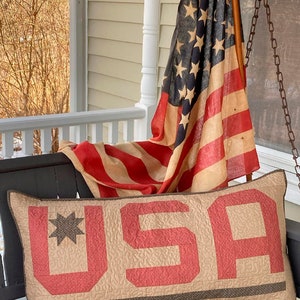 Op de afbeelding: Een gestikt kussen in tan en rood met het woord "USA" in grote rode letters. Het kussen ligt op een zwarte bank met een Amerikaanse vlag die over de rugleuning is gelegd.