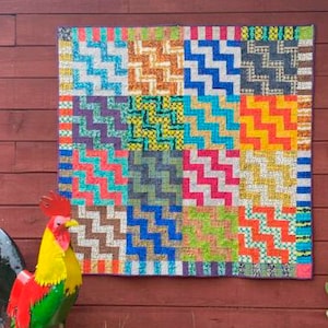 May include: A colourful patchwork quilt with a geometric pattern. The quilt is made up of squares of different colours and patterns, including red, blue, green, yellow, and orange. The quilt is hanging on a red wooden wall.