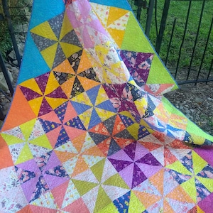 May include: A colourful quilt with a geometric pattern of pinwheels in shades of orange, yellow, green, purple, blue, and pink. The quilt is made of fabric with a floral print and is folded in half.