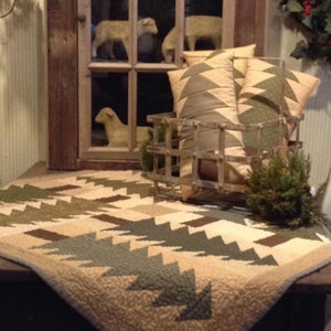 May include: A quilted throw featuring a pattern of green and brown stylized pine trees on a cream background. The throw is displayed on a wooden table with a metal basket and pillows in the background.