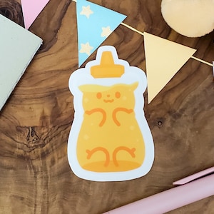 May include: A yellow honey bear sticker with a white outline. The bear is sitting in a honey jar with a white lid. The sticker is on a wooden surface.