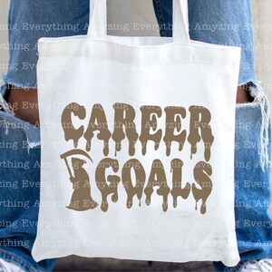 Puede incluir: Una bolsa de tela blanca con un gr&aacute;fico marr&oacute;n que dice "CAREER GOALS" con letras que gotean. El gr&aacute;fico tambi&eacute;n incluye una imagen estilizada de la Muerte.