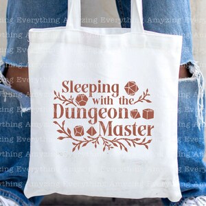May include: White tote bag with a brown design that says "Sleeping with the Dungeon Master". The design includes stylized dice and vines.