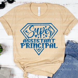 K&ouml;nnte beinhalten: Ein beiges T-Shirt mit einer blauen rautenf&ouml;rmigen Grafik, auf der "Super Assistant Principal" mit kleinen Herzen um den Text steht.