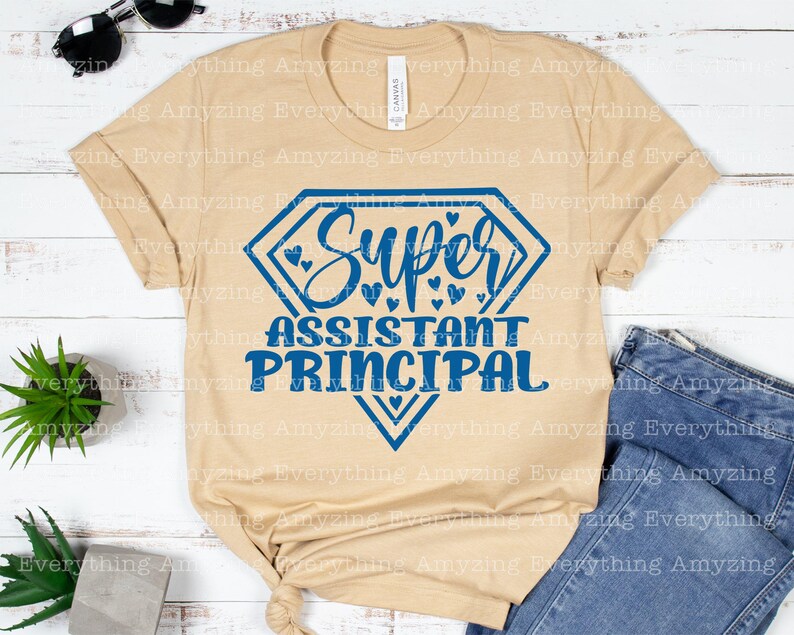 K&ouml;nnte beinhalten: Ein beiges T-Shirt mit einer blauen rautenf&ouml;rmigen Grafik, auf der "Super Assistant Principal" mit kleinen Herzen um den Text steht.