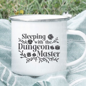 May include: White enamel mug with a black design that says "Sleeping with the Dungeon Master" and features several dice. The mug has a black rim and handle.