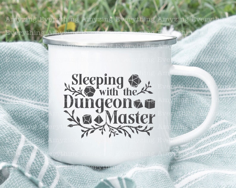 May include: White enamel mug with a black design that says "Sleeping with the Dungeon Master" and features several dice. The mug has a black rim and handle.