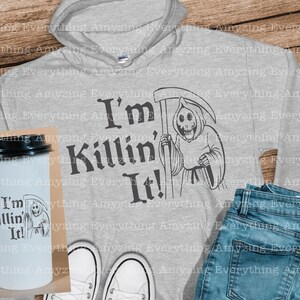 May include: A gray hoodie with a black graphic of a skeleton holding a scythe and the text "I'm Killin' It!"  A white coffee mug with the same graphic is also shown.