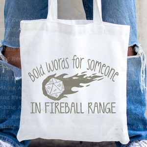 May include: White canvas tote bag with a gray graphic of a 20-sided die on fire and the text "Bold words for someone in fireball range".