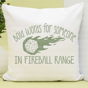 May include: White pillow with green text that reads "Bold words for someone in fireball range". A green 20-sided die with flames behind it is in the center of the pillow.