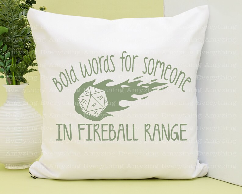 May include: White pillow with green text that reads "Bold words for someone in fireball range". A green 20-sided die with flames behind it is in the center of the pillow.